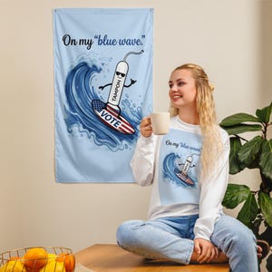 Può includere: Una maglietta bianca a maniche lunghe e uno stendardo da parete abbinato con un tampone a cartone animato che indossa occhiali da sole e fa surf su un'onda. La tavola da surf è decorata con la bandiera americana e la parola VOTE. Il testo dice On my blue wave.