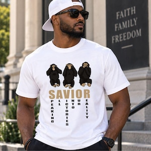 Puede incluir: Una camiseta blanca de manga corta con un gráfico de tres chimpancés cubriéndose los ojos, los oídos y la boca. Debajo de los chimpancés, la palabra SAVIOR está impresa en dorado, con las palabras PEAKING, ALL, VICTORIES, INTO, OUR, REALITY.