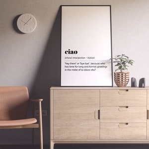 May include: A white framed poster with the word "ciao" in black text. The poster defines "ciao" as an Italian interjection meaning "hey there" or "bye bye". The poster is hanging on a light grey wall above a wooden cabinet with three drawers. A potted plant with green leaves and a copper-coloured pot is on the cabinet.