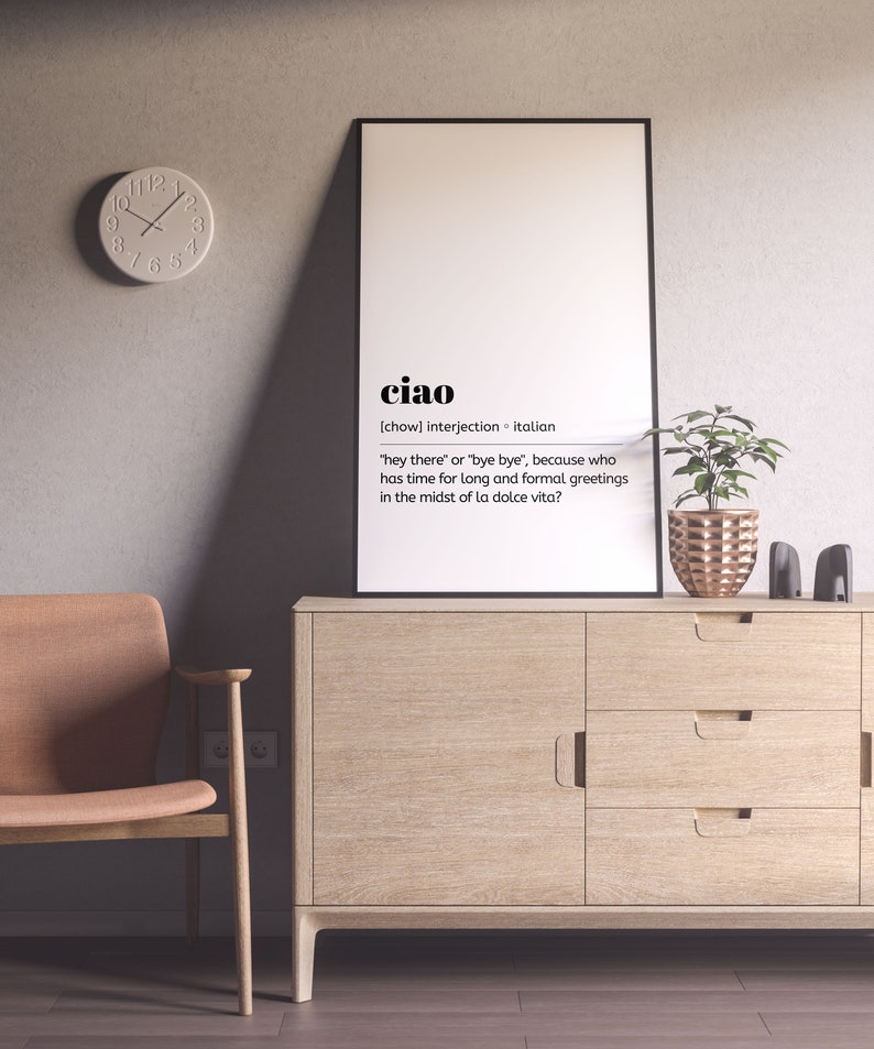 May include: A white framed poster with the word "ciao" in black text. The poster defines "ciao" as an Italian interjection meaning "hey there" or "bye bye". The poster is hanging on a light grey wall above a wooden cabinet with three drawers. A potted plant with green leaves and a copper-coloured pot is on the cabinet.