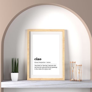 May include: A framed print with a white background and black text that reads "ciao" and its definition in Italian. The print is hanging on a wall with a light brown archway above it. There is a small potted plant and two hourglass sand timers on a white marble shelf below the print.
