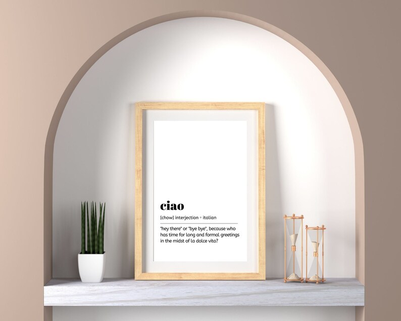 May include: A framed print with a white background and black text that reads "ciao" and its definition in Italian. The print is hanging on a wall with a light brown archway above it. There is a small potted plant and two hourglass sand timers on a white marble shelf below the print.