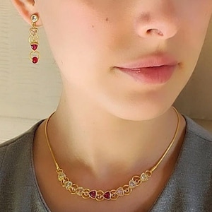 Puede incluir: Un collar de oro con una cadena delicada y una fila de piedras en forma de corazón alternando transparentes y rojas. El collar lo lleva una persona con una camisa gris.