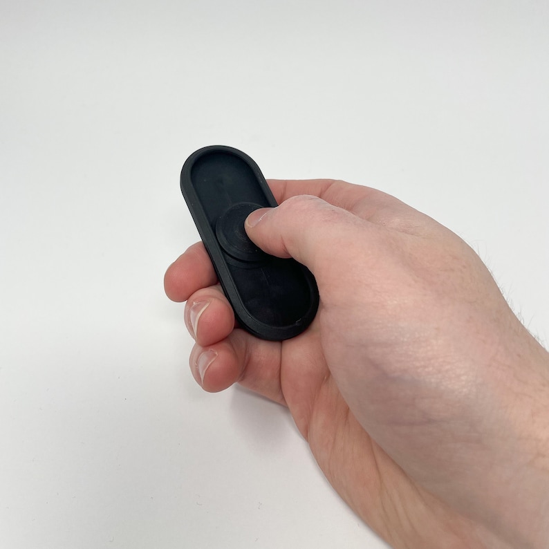 Puede incluir: Un fidget spinner negro se sostiene en una mano sobre un fondo blanco. El spinner tiene una forma rectangular redondeada con un rodamiento circular en el centro. Los dedos de la persona presionan el rodamiento central.