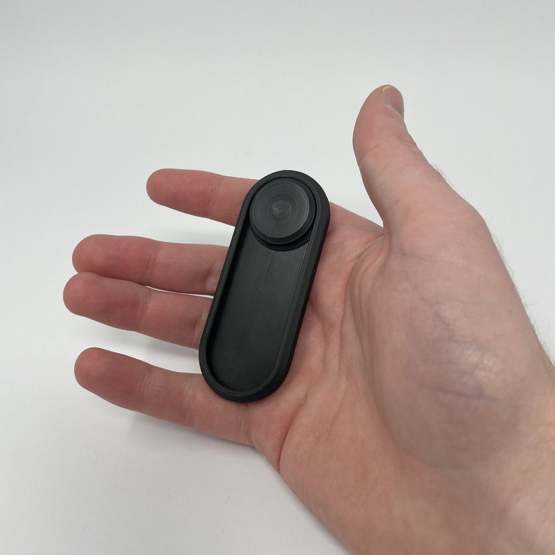 Puede incluir: Un objeto negro de forma oblonga con una parte superior circular, sostenido en una mano. El objeto parece estar hecho de un material mate. El dise&ntilde;o es simple y moderno, con un enfoque en la funcionalidad.