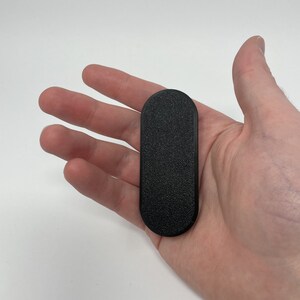 Puede incluir: Un soporte para tel&eacute;fono rectangular negro y texturizado con extremos redondeados. El soporte se sostiene en una mano sobre un fondo blanco. El soporte parece estar hecho de pl&aacute;stico o un material similar.