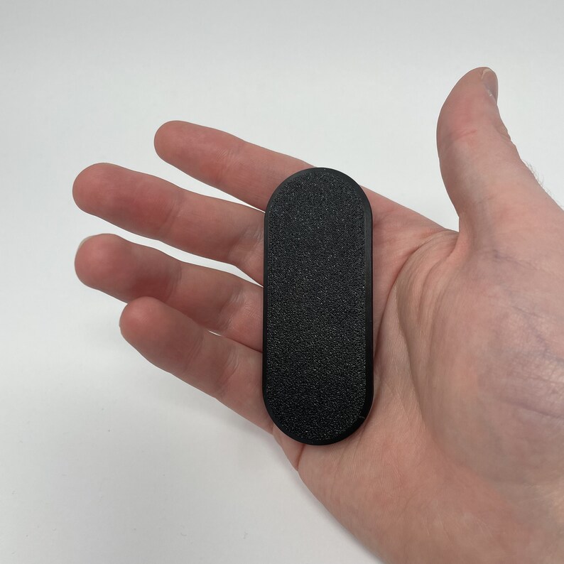Puede incluir: Un soporte para tel&eacute;fono rectangular negro y texturizado con extremos redondeados. El soporte se sostiene en una mano sobre un fondo blanco. El soporte parece estar hecho de pl&aacute;stico o un material similar.