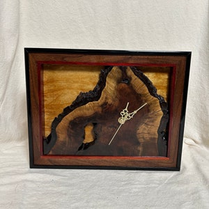 May include: A rectangular wooden clock with a black frame, featuring a natural wood grain face. The clock face has a dark brown, organic shape with a lighter wood background and red accents. Gold-colored clock hands complete the design.