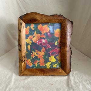 May include: A wooden picture frame with a natural, rustic edge. The frame holds a colorful image of orange, yellow, and red flowers. The wood grain is visible, adding to the frame's unique, handcrafted appearance.