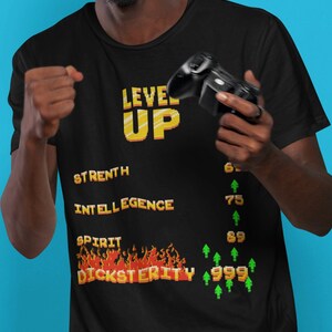Könnte beinhalten: Schwarzes T-Shirt mit Retro-Videospiel-Design. Das Shirt zeigt die Worte "LEVEL UP" in gelber Pixel-Schrift sowie "STRENTH", "INTELLIGENCE", "SPIRIT" und "DICKSTERITY". Das Shirt zeigt auch eine Reihe von Zahlen und grünen Pfeilen.
