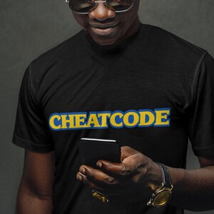 May include: A person wearing a black t-shirt with the text "CHEATCODE" in yellow and blue letters. The person is holding a black smartphone.