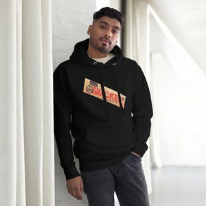 Op de afbeelding: Zwarte hoodie met een grafisch ontwerp met het woord "Smacke" in rood en wit op een bruine achtergrond. De hoodie heeft een capuchon met trekkoord en een voorzak. De persoon draagt een wit ondershirt en een donkere jeans.