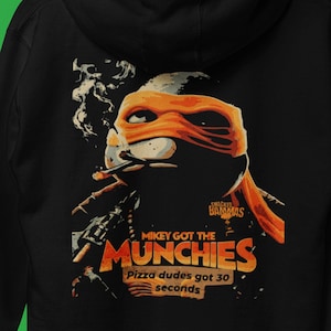 May include: A black hoodie featuring a graphic print of a cartoon ninja turtle smoking a cigarette. The graphic reads "Mikey got the munchies, Pizza dudes got 30 seconds" and "Snacked Bammas".