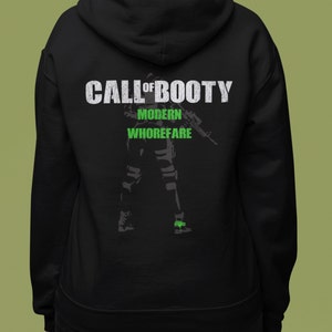 Pode incluir: Camisola de moletom com capuz preta com uma estampa gráfica verde de um soldado segurando um rifle. O texto "Call of Booty" é impresso acima do soldado, e o texto "Modern Whorefare" é impresso abaixo do soldado.