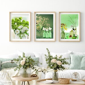 May include: Three framed prints with green and white floral arrangements and green glass bottles. The prints are hanging on a white wall above a white couch with green throw pillows. There are two small potted plants on the couch and a wooden coffee table in front of the couch.