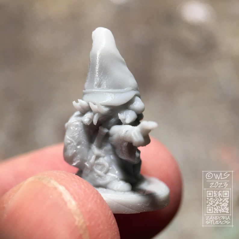 Pu&ograve; includere: Una miniatura di gnomo stampata in 3D di colore grigio, che indossa un cappello a punta e tiene un bastone. Lo gnomo indossa un mantello e ha la barba. La miniatura &egrave; su una base rotonda.