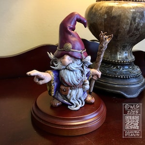 Pu&ograve; includere: Una statuetta dipinta a mano di un mago con una lunga barba bianca, che indossa una tunica viola e un cappello bordeaux con una fascia dorata. Tiene in mano un bastone di legno con una cima a forma di albero e indica con la mano sinistra. La statuetta &egrave; su una base di legno.