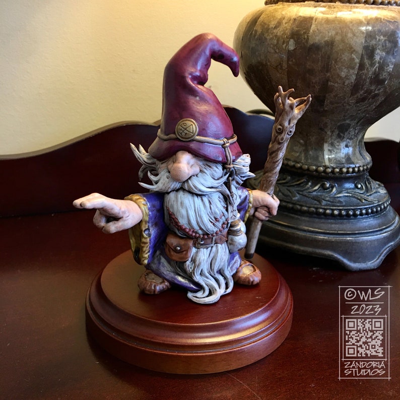 Pu&ograve; includere: Una statuetta dipinta a mano di un mago con una lunga barba bianca, che indossa una tunica viola e un cappello bordeaux con una fascia dorata. Tiene in mano un bastone di legno con una cima a forma di albero e indica con la mano sinistra. La statuetta &egrave; su una base di legno.