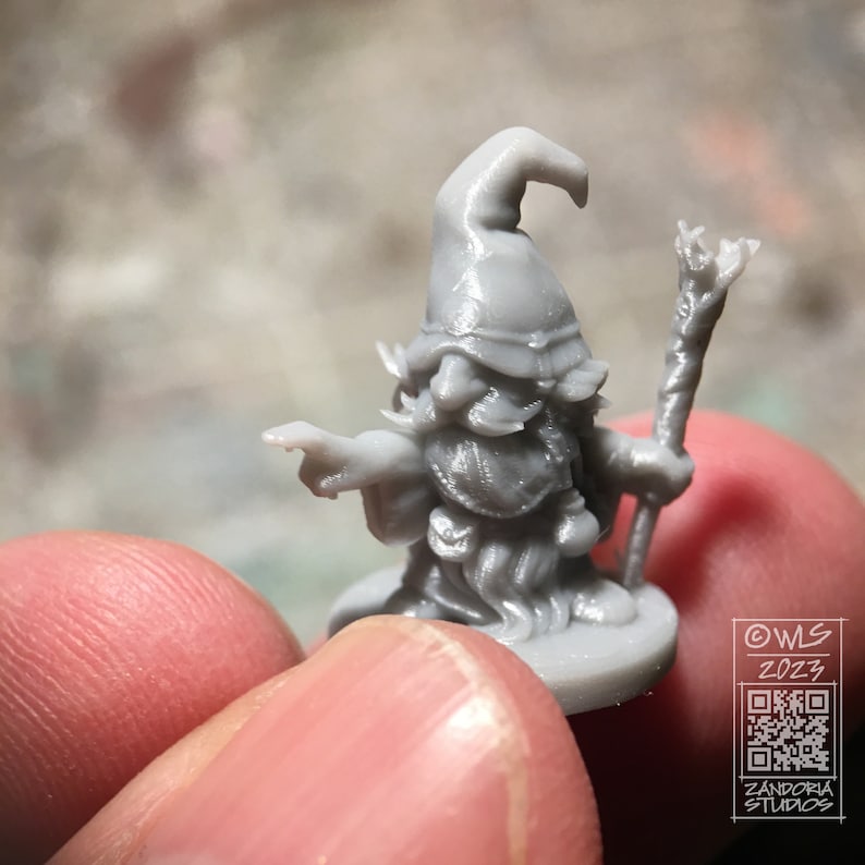Pu&ograve; includere: Una miniatura grigia di un mago stampata in 3D con un cappello a punta, barba e bastone. La miniatura &egrave; in piedi su una base rotonda.