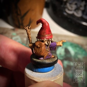 Pu&ograve; includere: Una statuetta in miniatura dipinta di un gnomo che indossa un cappello rosso, una tunica viola e una borsa marrone. Lo gnomo tiene un bastone e indica con una mano. La statuetta &egrave; su una base rotonda con un anello blu.