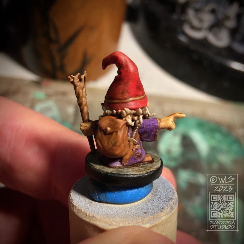 Pu&ograve; includere: Una statuetta in miniatura dipinta di un gnomo che indossa un cappello rosso, una tunica viola e una borsa marrone. Lo gnomo tiene un bastone e indica con una mano. La statuetta &egrave; su una base rotonda con un anello blu.