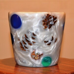 May include: White ceramic cup with blue, green and brown accents. The cup has a swirl pattern and is decorated with pine cones.