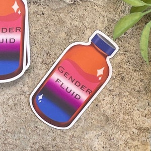 May include: A set of stickers featuring a cartoon illustration of a bottle with the text "Gender Fluid". The bottle is orange with a pink, white, and blue liquid inside. The stickers are on a brown textured surface.