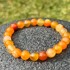 May include: A bracelet made of orange and yellow agate beads. The round, polished beads vary in shade from orange to yellow and white. The bracelet is displayed on a grey, textured surface, with a blurred green background.