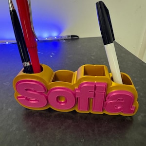 Puede incluir: Organizador de escritorio personalizado en oro y rosa, con el nombre "Sofia" en letras rosas en relieve. Contiene un bolígrafo negro, un bolígrafo rojo y un rotulador negro. Diseño impreso en 3D.