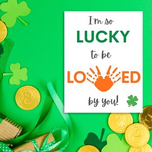 May include: A white card with the text "I'm so LUCKY to be LOVED by you!" with a handprint in orange. The card is surrounded by green shamrocks and gold coins.