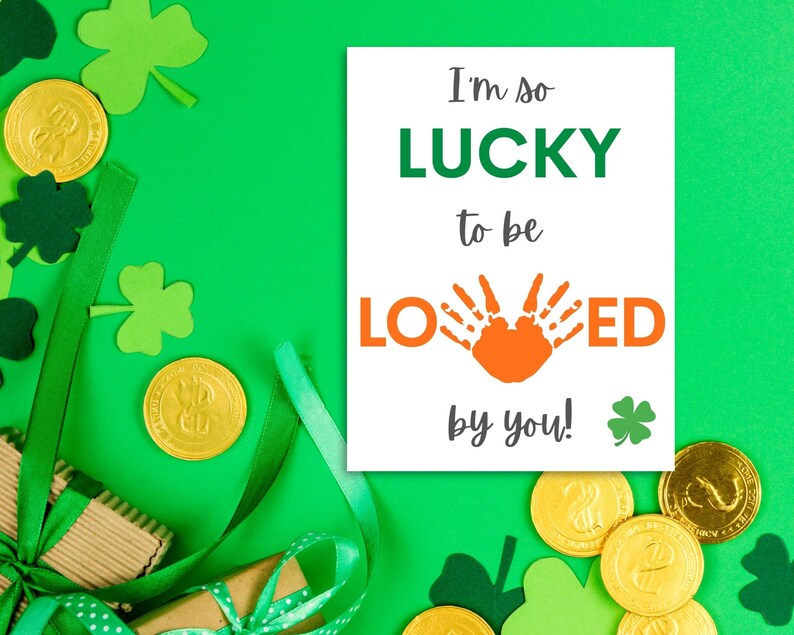 May include: A white card with the text "I'm so LUCKY to be LOVED by you!" with a handprint in orange. The card is surrounded by green shamrocks and gold coins.