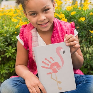 May include: A young person is holding a white card with a pink flamingo handprint design. The flamingo is outlined in black and has a pink beak. The card is against a background of yellow flowers.
