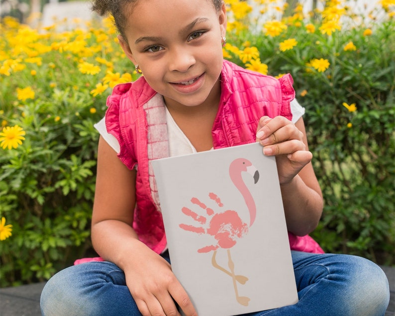 May include: A young person is holding a white card with a pink flamingo handprint design. The flamingo is outlined in black and has a pink beak. The card is against a background of yellow flowers.