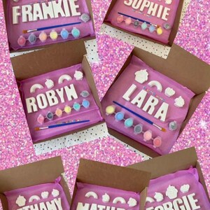May include: Six paint-your-own name kits in pink boxes. Each kit includes a set of white ceramic letters spelling a name, paint pots, a paintbrush, and colorful sprinkles. The names are Frankie, Sophie, Robyn, Lara, Bethany, and Matilda.