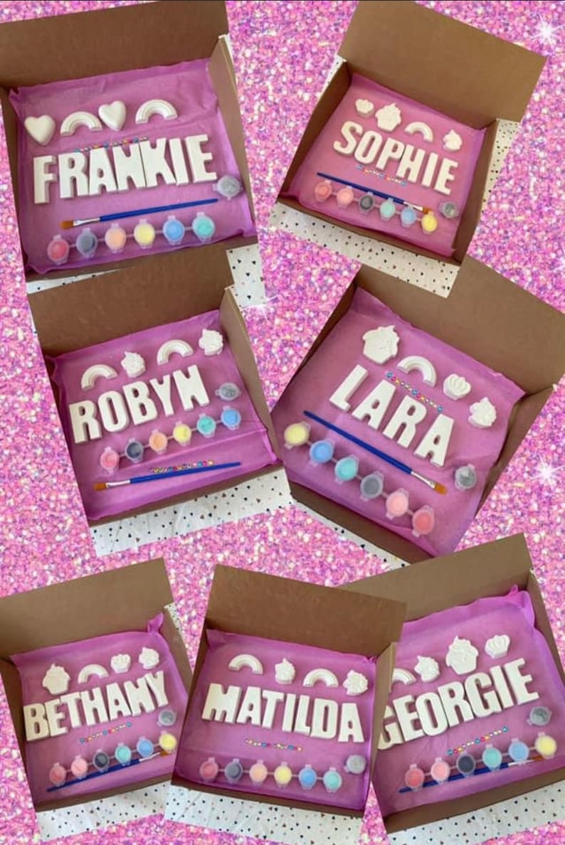 May include: Six paint-your-own name kits in pink boxes. Each kit includes a set of white ceramic letters spelling a name, paint pots, a paintbrush, and colorful sprinkles. The names are Frankie, Sophie, Robyn, Lara, Bethany, and Matilda.