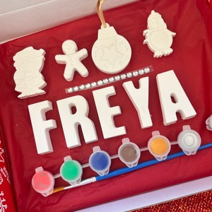 May include: A Christmas craft kit featuring white ceramic ornaments, including a snowman, gingerbread person, bauble, and the name "FREYA" in large letters. The kit comes with paints, a brush, and glitter.