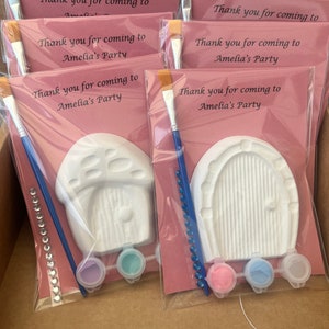 May include: Four pink and white favor bags with paint sets for a party. Each bag includes a paintbrush, two small paint pots, and a white ceramic figurine. The figurines are a mushroom house and a door. The bags have the text "Thank you for coming to Amelia's Party" printed on them.