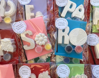 May include: Assortment of party favors, each individually packaged. The favors include white, paintable shapes like letters, a flower, a rocket, and a soccer ball, along with small paint pots. Each package has a sticker that says "Thank you for coming to Albie's Party."