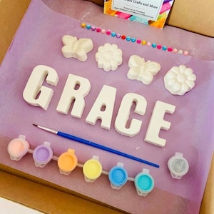 May include: A craft kit for painting a name sign. The kit includes the letters G-R-A-C-E, two butterflies, two flowers, a paintbrush, and eight small paint pots in various pastel colors. The kit is packaged in a cardboard box with a pink and purple tissue paper lining. The box also includes a business card for Letterbox Crafts and More.
