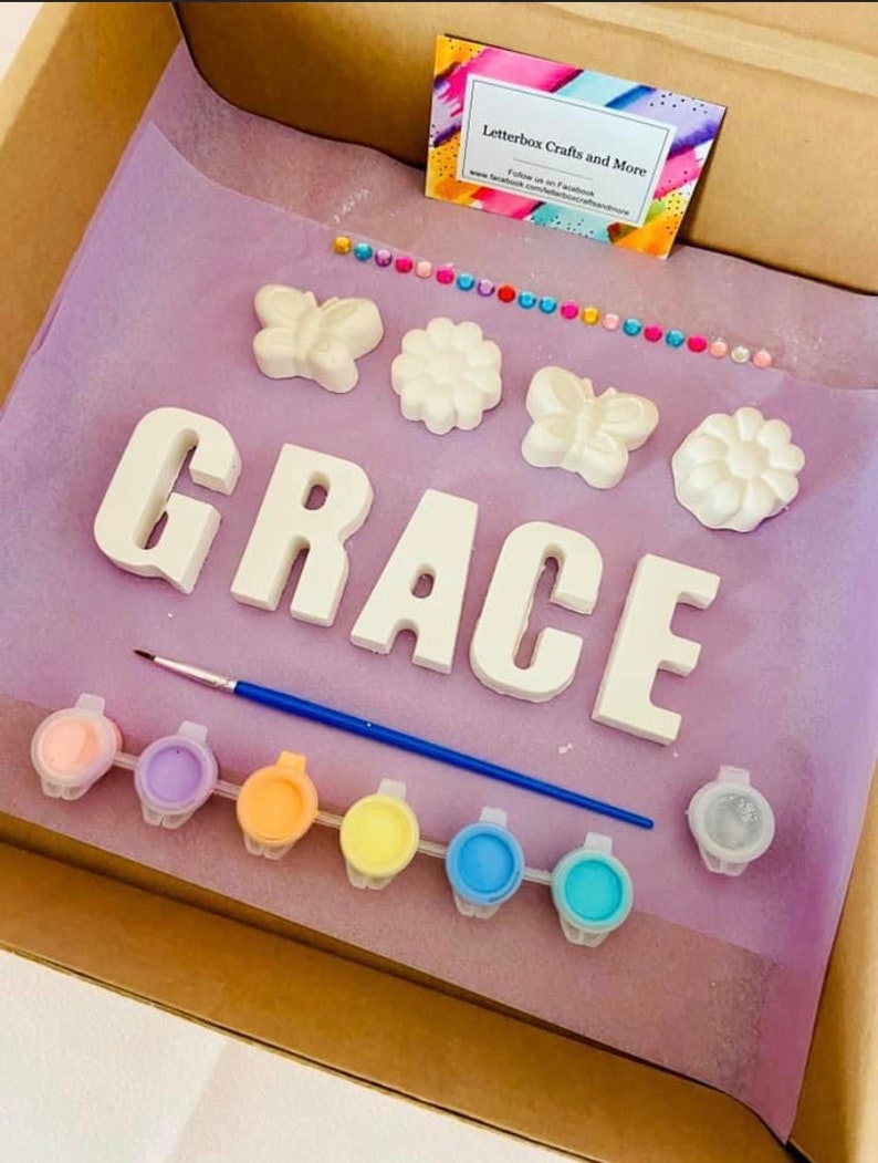 May include: A craft kit for painting a name sign. The kit includes the letters G-R-A-C-E, two butterflies, two flowers, a paintbrush, and eight small paint pots in various pastel colors. The kit is packaged in a cardboard box with a pink and purple tissue paper lining. The box also includes a business card for Letterbox Crafts and More.