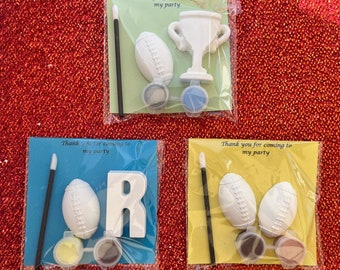 May include: Three sets of paint your own party favors. Each set includes a small paint brush, two paint pots, and a ceramic figure. The figures are a football, a trophy, and the letter R. The background is a red sparkly surface. The text on the cards reads "Thank you for coming to my party."