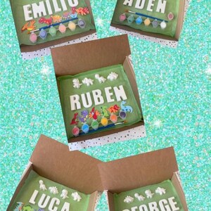 May include: Four open cardboard boxes each contain a paint your own dinosaur craft kit. Each kit includes a set of ceramic dinosaur figurines, a paintbrush, and a set of acrylic paints. The names "Emilio", "Aiden", "Ruben", "Luca", and "George" are spelled out in ceramic letters in each box.