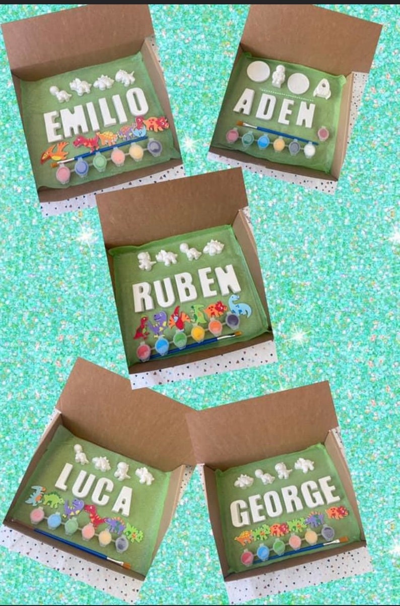 May include: Four open cardboard boxes each contain a paint your own dinosaur craft kit. Each kit includes a set of ceramic dinosaur figurines, a paintbrush, and a set of acrylic paints. The names "Emilio", "Aiden", "Ruben", "Luca", and "George" are spelled out in ceramic letters in each box.