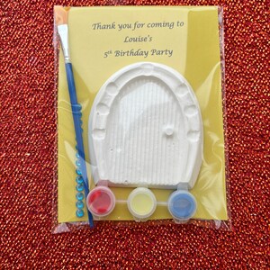 May include: A craft kit for a birthday party with a white plaster door, red, yellow, and blue paint, a paintbrush, and blue rhinestones. The kit has a label that says "Thank you for coming to Louise's 5th Birthday Party."