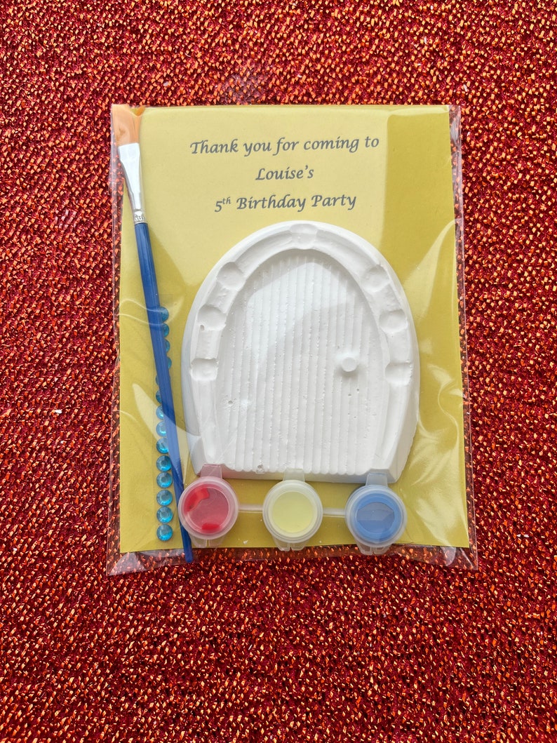 May include: A craft kit for a birthday party with a white plaster door, red, yellow, and blue paint, a paintbrush, and blue rhinestones. The kit has a label that says "Thank you for coming to Louise's 5th Birthday Party."