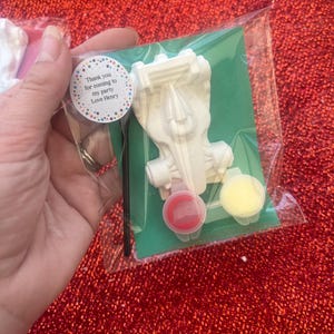 May include: A clear plastic bag contains a white, unpainted toy spaceship, two small paint pots with red and yellow paint, a paintbrush, and a sticker that reads "Thank you for coming to my party Love Henry". The contents rest on a green card.