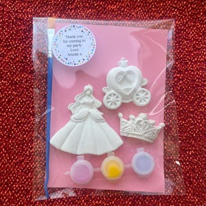 May include: A craft kit in a clear plastic bag containing three white plaster figures: a princess, a carriage, and a crown. It includes a blue paintbrush and three small paint pots in pink, yellow, and purple. A round sticker reads: Thank you for coming to my party Love Amelie x.