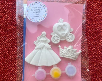 Pode incluir: Um kit de artesanato num saco de plástico transparente contendo três figuras de gesso branco: uma princesa, uma carruagem e uma coroa. Inclui um pincel azul e três pequenos potes de tinta rosa, amarela e roxa. Um autocolante redondo diz: Thank you for coming to my party Love Amelie x.