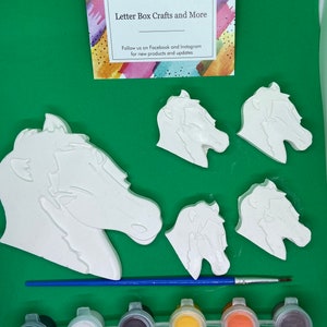 May include: A craft kit featuring five white ceramic horse head figurines and six paint pots. The paint pots are black, white, brown, yellow, orange, and purple. The kit also includes a blue paintbrush. The card in the image reads "Letter Box Crafts and More" and "Follow us on Facebook and Instagram for new products and updates."