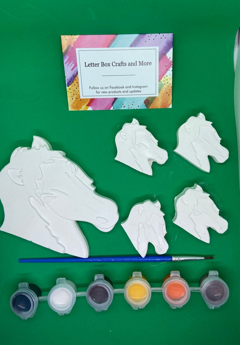 May include: A craft kit featuring five white ceramic horse head figurines and six paint pots. The paint pots are black, white, brown, yellow, orange, and purple. The kit also includes a blue paintbrush. The card in the image reads "Letter Box Crafts and More" and "Follow us on Facebook and Instagram for new products and updates."